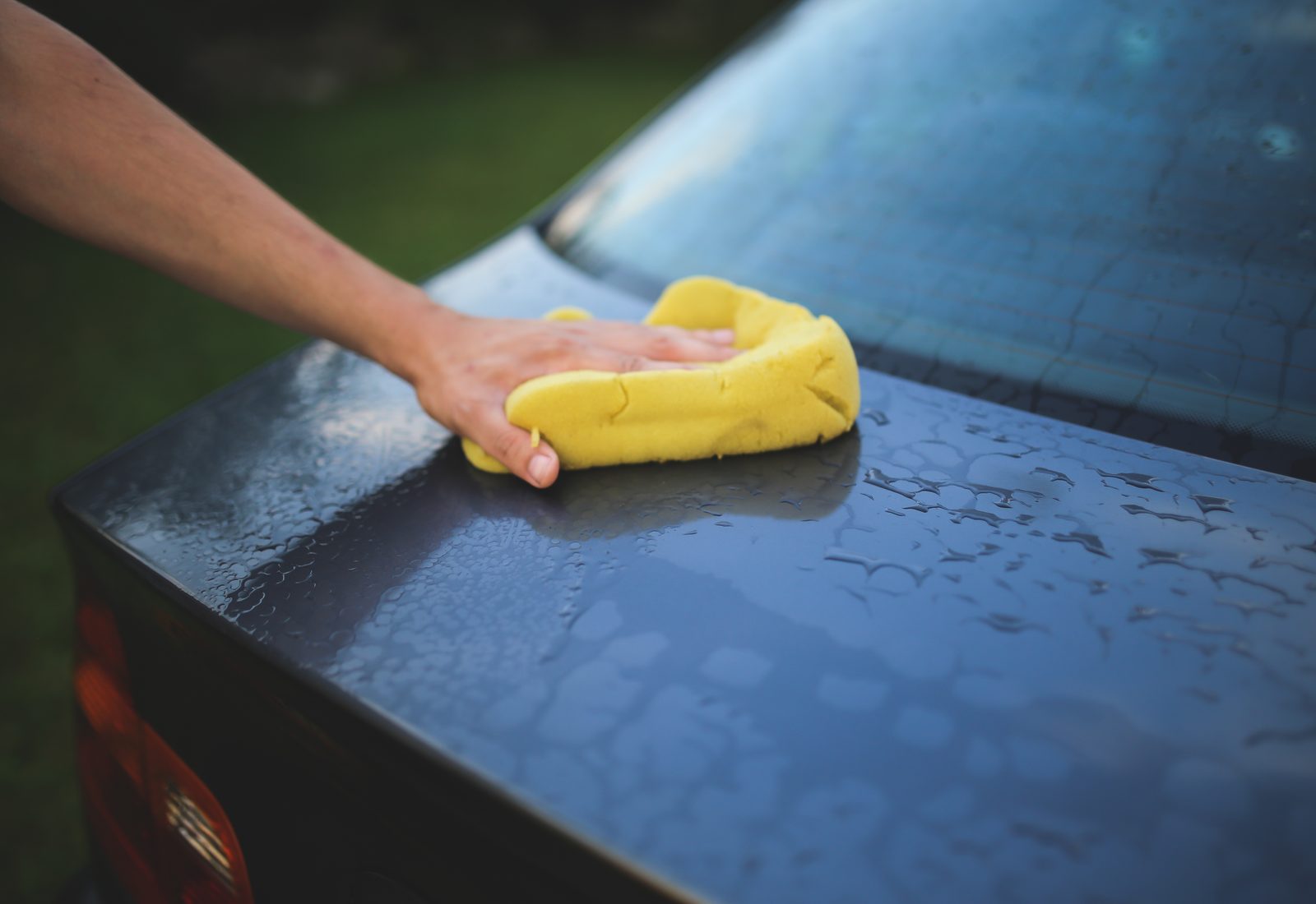 A hand washing a dark car with a soapy yellow sponge